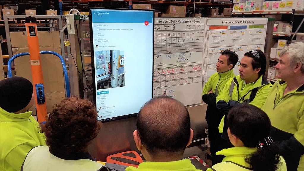 A shiftly tiered meeting in a manufacturing site, with a digital primary visual display and whiteboards.